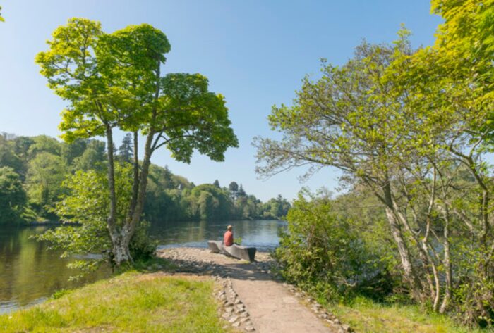 A person in red sits on a quiet bench on the shores of the River Ness.